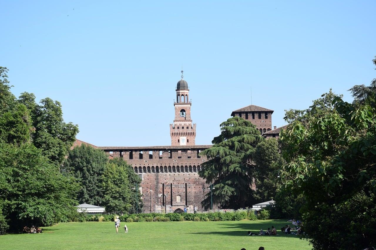 Il Castello Sforzesco di Milano con la torre in mattoni, visto da un prato verde del parco con alberi e persone sdraiate.